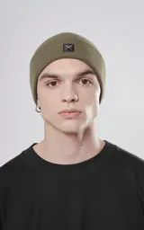 Gorro de tejido de punto color verde militar, de estilo beanie, con borde doblado y un pequeño parche negro con un logo de tijeras cruzadas.