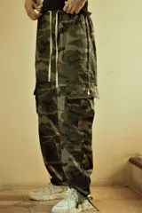 Pantalón cargo holgado con estampado camuflado en tonos verde militar, marrón y negro. Presenta cintura elástica ajustable con cordón blanco y bolsillos cargo laterales con cierre de botón a presión. Los tobillos son ajustables con cordones.