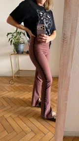 Pantalón de gabardina elastizada color malva o rosado oscuro, de tiro alto y corte acampanado. Presenta un ribete decorativo negro con borde festoneado a lo largo de las costuras laterales exteriores de las piernas.