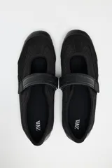 Zapato tipo bailarina color negro, con capellada de cuero sintético y rejilla. Tiene tira con velcro en el empeine y suela plana.
