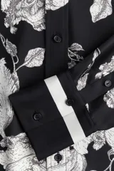 Camisa de sarga estampada con flores en blanco y negro, de corte recto, con cuello clásico, cierre frontal con botones, mangas largas con puños abotonados y bajo redondeado.