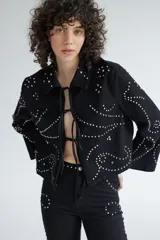 Chaqueta corta de denim negro con cuello clásico y cierre frontal con cordones. Decorada con tachas plateadas formando patrones ondulados.