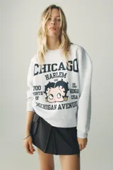 Sudadera gris melange de cuello redondo y manga larga con puños elásticos. Estampado frontal con texto "Chicago Harlem" y dibujo de Betty Boop.
