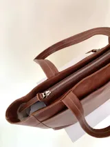 Cartera tipo tote de cuero graneado color negro, con textura uniforme y suave al tacto. Tiene manijas de 55 cm, bolsillo frontal con broche imantado, bolsillo trasero con cierre metálico, cierre principal metálico, forro interior y tres bolsillos internos (uno con cierre, uno plaqué y uno tipo sobre con solapa y botón).