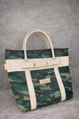 Bolso de playa tipo tote bag con estampado camuflado en tonos verdes y beige. Tiene doble asa de hombro color beige y detalles en cuero marrón en el borde superior. Incluye una tira horizontal con hebilla decorativa y un parche con el logo de la marca SierraMora.