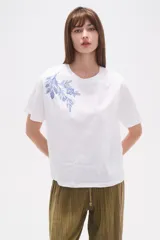 Remera blanca de manga corta y cuello redondo, con un bordado floral en tonos azules en la zona superior izquierda del pecho.