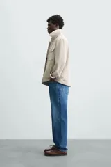 Sobrecamisa color beige de corte relaxed fit, confeccionada en tejido con elasticidad. Tiene cuello solapa, manga larga con puños abotonados, bolsillos de parche con solapa en el pecho y bolsillos de ojal en la cadera. El cierre es frontal con botones.