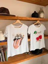 Remera blanca de algodón con cuello redondo y estampado de cerezas rojas con hojas verdes y texto dorado.