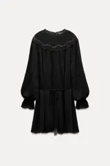 Vestido corto negro con cuello redondo, mangas largas abullonadas con puños elásticos y detalle de encaje y bordados con abalorios. Incluye lazo para ajustar en la cintura.