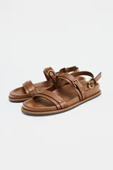Sandalias planas color marrón con doble tira delantera con detalle de hebilla y cierre mediante hebilla en la pulsera al tobillo.
