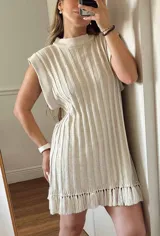 Vestido corto color beige tejido con hilo de algodón y lurex en punto acanalado vertical y flecos. Tiene corte recto, cuello redondo con una guarda ancha y hombros amplios que dan efecto de hombrera caída.