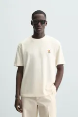 Camiseta color crudo de corte relaxed fit, con cuello redondo y mangas cortas. Presenta un pequeño bordado de flores en el pecho y un estampado fotográfico en la espalda.