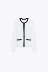 Chaqueta blanca de punto con cuello redondo y manga larga. Tiene falsos bolsillos de vivo en el delantero y cierre frontal con botones dorados.