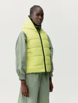 Chaleco puffer reversible con capucha, color amarillo de un lado y gris del otro. Cierre frontal con cremallera de doble cursor y bolsillos laterales con cremallera. Tejido repelente al viento y relleno 100% vegano.