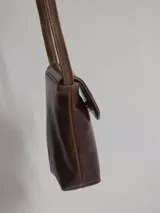 Bandolera porta celular marrón de cuero con solapa y cierre con botón a presión. Presenta tachas metálicas plateadas en el borde de la solapa y correa fina para colgar al hombro.
