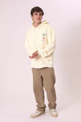 Sudadera con capucha color crema o amarillo muy pálido, de corte holgado, con bolsillo canguro frontal y cordones ajustables en la capucha. Presenta un pequeño estampado gráfico en el pecho con un cuadrado verde y una figura roja con texto debajo.