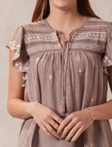 Blusa color marrón claro de algodón con bordados y calados blancos. Tiene cuello a la base, escote en V con lazo de ajuste y mangas cortas amplias.