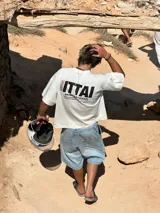 Remera color crema de corte recto y mangas cortas, con estampado en la espalda con el logo "ITTAI" en negro con líneas rojas, azules y blancas, y la leyenda "AN IBIZA PRODUCE. Ittai Stars Department Gallery - Ibiza Edition. If you're in Ibiza, 112 is not an option.".