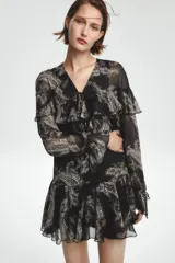 Vestido corto negro con estampado paisley en tonos beige y detalles dorados. Escote en V con lazos ajustables, mangas largas con puños elásticos y volantes en el mismo tejido en el escote y el ruedo.