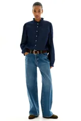 Camisa de denim azul oscuro con cuello mao y volados en cuello y puños. Tiene mangas largas abullonadas con botón en los puños, botonadura frontal y bajo redondeado.