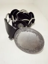 Cinto negro de cuero con argollas plateadas y hebilla metálica ovalada con diseño floral repujado.