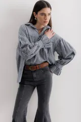 Camisa celeste a rayas verticales blancas, de corte holgado, con cuello camisero y mangas largas abullonadas con puños elásticos.