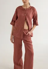 Conjunto de chaqueta y pantalón color ladrillo, confeccionado en lino. La chaqueta es de corte holgado, con escote redondo, mangas 3/4 y cierre frontal con botones de madera. Tiene bolsillos de parche con costuras a contraste. El pantalón es de corte recto y cintura elástica.