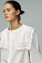 Camisa blanca de popelín de algodón con cuello redondo con tejido en superposición a tono y aplicación de bordados. Manga larga acabada en elástico y cierre frontal con botones ocultos por sopata.