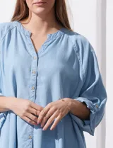 Camisa celeste de tencel, corte amplio, con cuello mao, escote en V y abotonadura frontal. Tiene bolsillos aplicados en el pecho, mangas 3/4 regulables con presilla y ruedo desflecado.