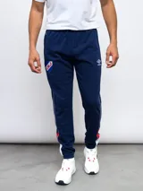 Pantalón de jogging azul marino con puños ajustados, cintura elástica con cordón y bolsillos laterales. Presenta una franja blanca con detalles en rojo a lo largo de la pierna, el escudo del Club Nacional de Football y el logo de Umbro.