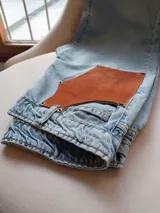 Pantalón de jean celeste con cintura elastizada, corte recto, bolsillos laterales y aplique de cuero marrón en el frente. Presenta botones metálicos en los laterales y etiqueta de cuero en la parte posterior.