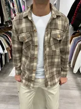 Sobrecamisa de paño color tostado con diseño a cuadros en tonos beige y marrón. Tiene cuello clásico, cierre frontal con botones de madera, dos bolsillos con solapa en el pecho y puños abotonados.