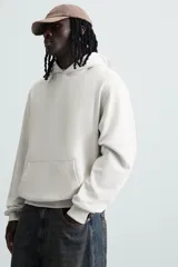Sudadera con capucha color crudo, de corte relajado, confeccionada en tejido de algodón con interior rizado. Cuenta con cuello con capucha, manga larga, bolsillo frontal tipo canguro y acabados en rib.