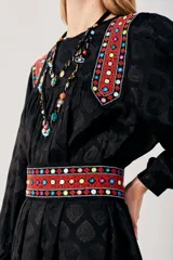 Vestido largo negro con estampado de hojas en relieve, mangas largas abullonadas y falda plisada. Presenta detalles bordados en rojo, blanco y azul en los hombros y la cintura.