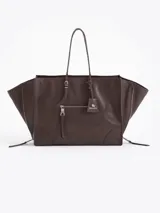 Bolso tote de cuero vacuno color marrón oscuro, con forma trapezoidal y asas de hombro. Presenta cierres funcionales en los laterales para expandir la capacidad y un bolsillo frontal con cierre y tirador metálico. Incluye un tag colgante con aplique metálico.