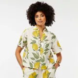 Blusa color crema con estampado de limones y hojas verdes, de manga corta, cuello camisero y cierre frontal con botones.