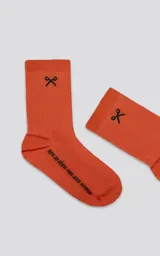 Medias 3/4 de algodón color naranja con textura acanalada. Presentan un bordado negro en la parte superior con un símbolo de tijeras y texto en la planta del pie que dice "ropa sin género para seres humanos".