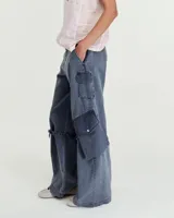 Pantalón cargo de gabardina gastada de lino y algodón color azul. Silueta de pierna recta ancha, con cartera con cierre metálico y broche níquel en el cruce de la cintura. Tiene bolsillos inclinados en los laterales y bolsillos cargo con tapa y broches metálicos a la altura de las rodillas, de los cuales salen unas tiras que se ajustan mediante hebillas metálicas para modificar la silueta del pantalón. En la espalda tiene pinzas y un bolsillo plaque con cierre metálico en boca. Es tiro medio.