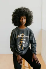 Buzo de manga larga color gris oscuro/negro desteñido, con cuello redondo y puños y ruedo acanalados. Estampado frontal con el logo de The Rolling Stones (lengua y labios) en dorado y texto en cursiva.