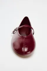 Zapato tipo bailarina color borgoña con efecto acharolado, acabado en punta redonda y tira con hebilla ajustable en la parte delantera.
