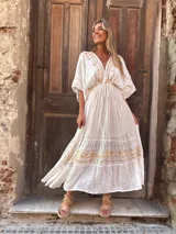 Vestido largo blanco con escote en V profundo y mangas cortas con volantes. Presenta detalles bordados o estampados en hilo dorado en el escote y en los volantes de la falda, con un ajuste fruncido en la cintura.