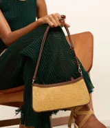 Cartera baguette color beige con efecto paja y detalles en cuero sintético marrón. Tiene correa de hombro ajustable y cierre por cremallera.