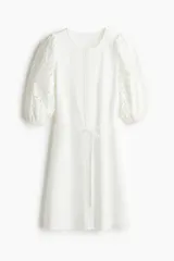 Vestido blanco a la rodilla en tejido liviano de algodón con cuello redondo, botones ocultos adelante y cinturón fino extraíble. Mangas al codo abullonadas en broderie con puños elásticos.