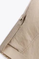 Camisa color beige de cuello solapa y manga corta con trabilla y botón. Tiene bolsillos delanteros de plastrón con solapa, bajo con elástico y cierre frontal con botones a presión.