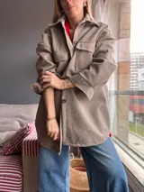 Sobre camisa color marrón claro, de corte holgado, con cuello camisero, cierre frontal con botones y bolsillos de parche en el pecho.