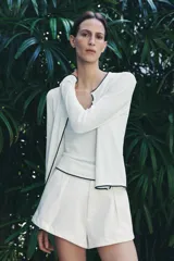 Chaqueta blanca de punto con cuello redondo y manga larga. Tiene vivos negros en el cuello, el borde frontal y los puños. Se cierra con botones en el frente.