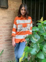 Camisa de manga 3/4 con cuello clásico y cierre frontal con botones. Diseño a rayas horizontales color naranja y blanco, con bolsillos frontales con solapa en color dorado.