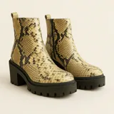 Bota de caña media con estampado de serpiente en tonos dorados y negros, cierre lateral y tiradores en la parte superior. Suela gruesa y robusta con tacón bajo.