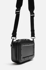 Bandolera rígida negra con formato de caja. Tiene un compartimento principal con cierre de cremallera, un bolsillo interior plano y asa de hombro ajustable y extraíble.