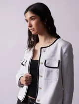 Chaqueta de tweed blanca con ribete negro, escote a la base, abertura central con botones, bolsillos delanteros y puños con aplique de botones perlados.